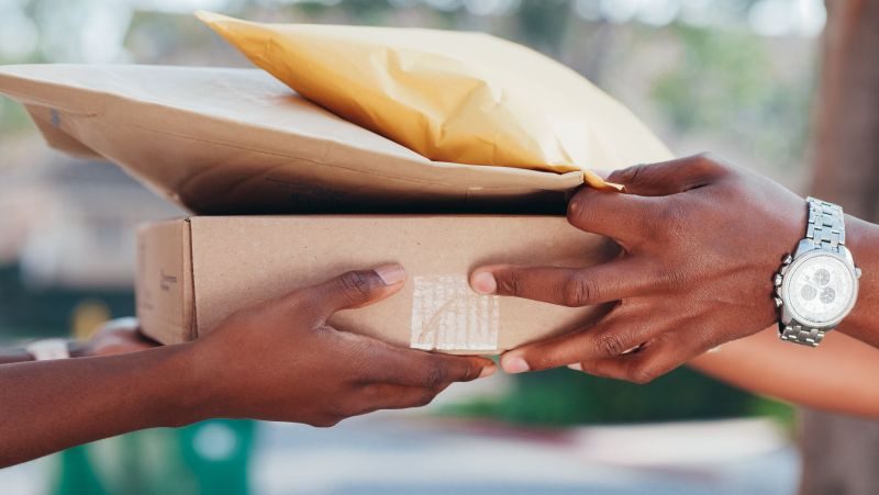 Two people exchange a cardboard parcel outdoors, hands meeting as the package passes between them, a wristwatch visible on one person.